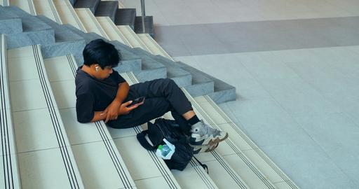 Young man sitting on indoor stairs using smartphone and wearing earbuds, resting