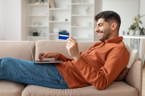 Smiling Young Man Using Laptop And Holding Credit Card At Home, Online Shopping