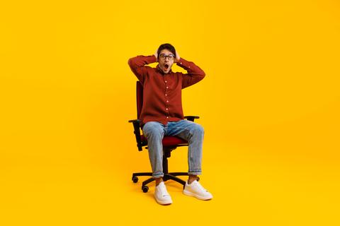 Surprised Young Man With Glasses Sitting In Office Chair, Yellow Background