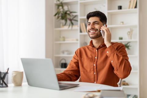 Smiling Young Man Talking On Phone At Desk With Laptop In Modern Office