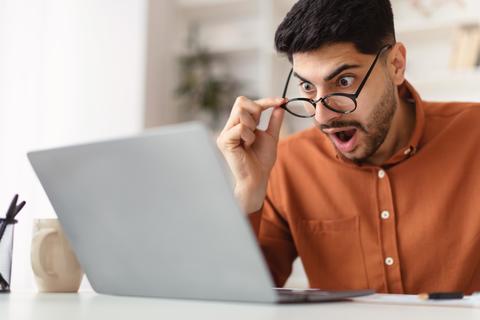 Shocked Young Man Wearing Glasses Staring At Laptop Screen In Home Office
