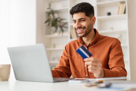 Smiling Young Man Holding Credit Card While Shopping Online At Laptop, Business