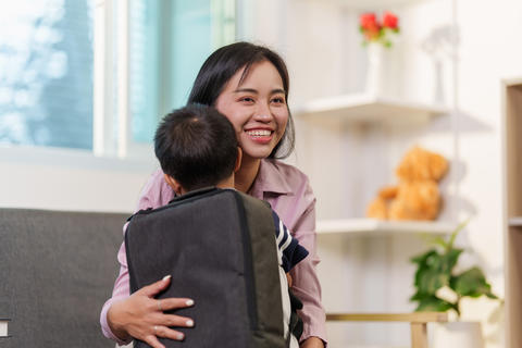 Happy Asian mother hugging her young son with backpack in cozy home living room