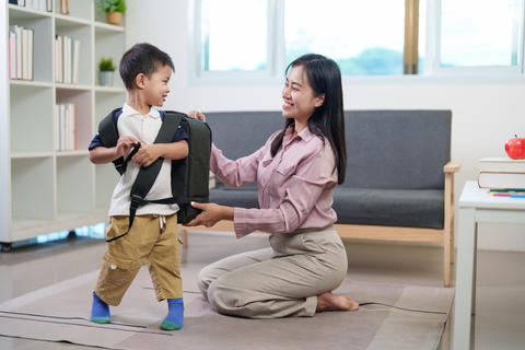 Asian mother helping young son wear backpack for first day of school at home