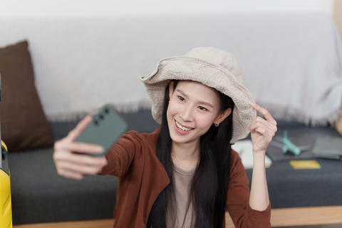 Smiling Asian woman wearing sun hat taking selfie with smartphone for travel