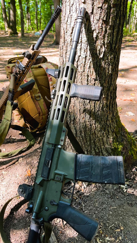 Military Rifle, Assault Rifle and Backpack Standing Near a Tree in a Forest