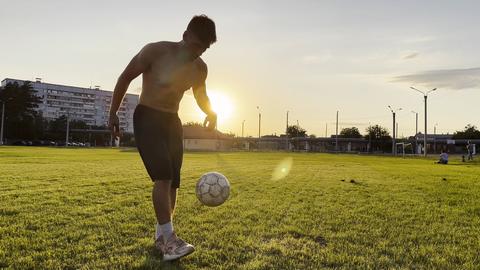 Young man juggling soccer ball on stadium at sunset. Professional footballer kicking ball at green f