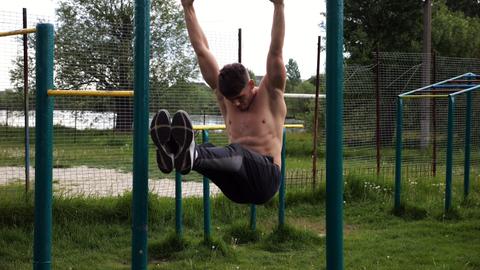 Athletic man trains abdominal muscles raising legs on horizontal bar at sports ground. Young sportsm