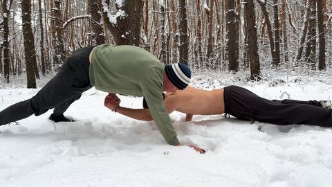 Two male athletes performing atypical partner push-ups on deep snow in winter forest. Strong sportsm